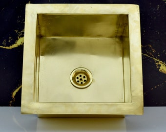 May include: A square brass sink with a drain in the center. The sink is set against a black background.