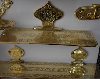 Puede incluir: Dos estantes de latón adornados con diseños intrincados, cada uno con un gancho de latón decorativo en el lateral. Los estantes están montados en una pared blanca.