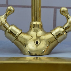 May include: A gold-toned double-handled faucet with a polished finish. The faucet has two separate handles, each with a round knob and a cross-shaped design. The faucet is mounted on a white tiled surface.