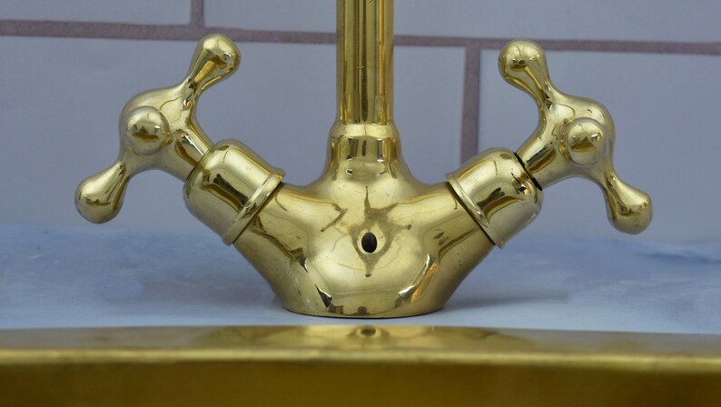 May include: A gold-toned double-handled faucet with a polished finish. The faucet has two separate handles, each with a round knob and a cross-shaped design. The faucet is mounted on a white tiled surface.