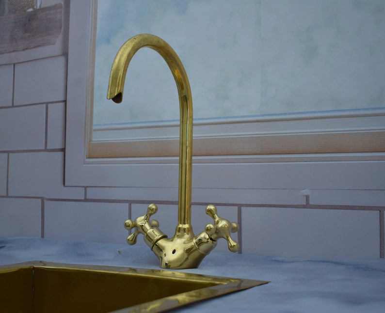 May include: A gold-toned faucet with two cross-shaped handles, mounted on a white tiled countertop. The faucet has a curved spout and is attached to a sink.