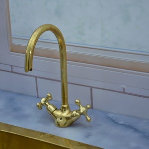 May include: A gold-toned kitchen faucet with a high-arc spout and two cross-shaped handles. The faucet is mounted on a white marble countertop.