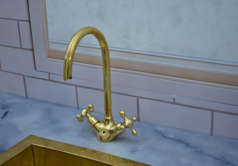 May include: A gold-toned kitchen faucet with a high-arc spout and two cross-shaped handles. The faucet is mounted on a white marble countertop.