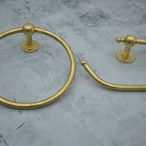May include: Two gold-toned towel rings with a textured finish. The larger ring is circular, while the smaller ring is a curved shape.