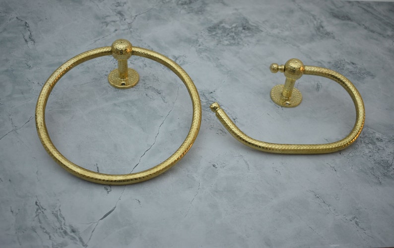 May include: Two gold-toned towel rings with a textured finish. The larger ring is circular, while the smaller ring is a curved shape.