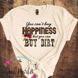 May include: A white t-shirt with the text "You can't buy happiness but you can buy dirt" printed on it. The text is in a brown and black camouflage pattern with pink flowers.