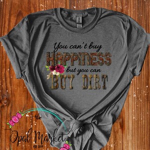 May include: Gray t-shirt with the text "You can't buy happiness but you can buy dirt" in brown and gold lettering with a floral design. The text is surrounded by a camouflage pattern.