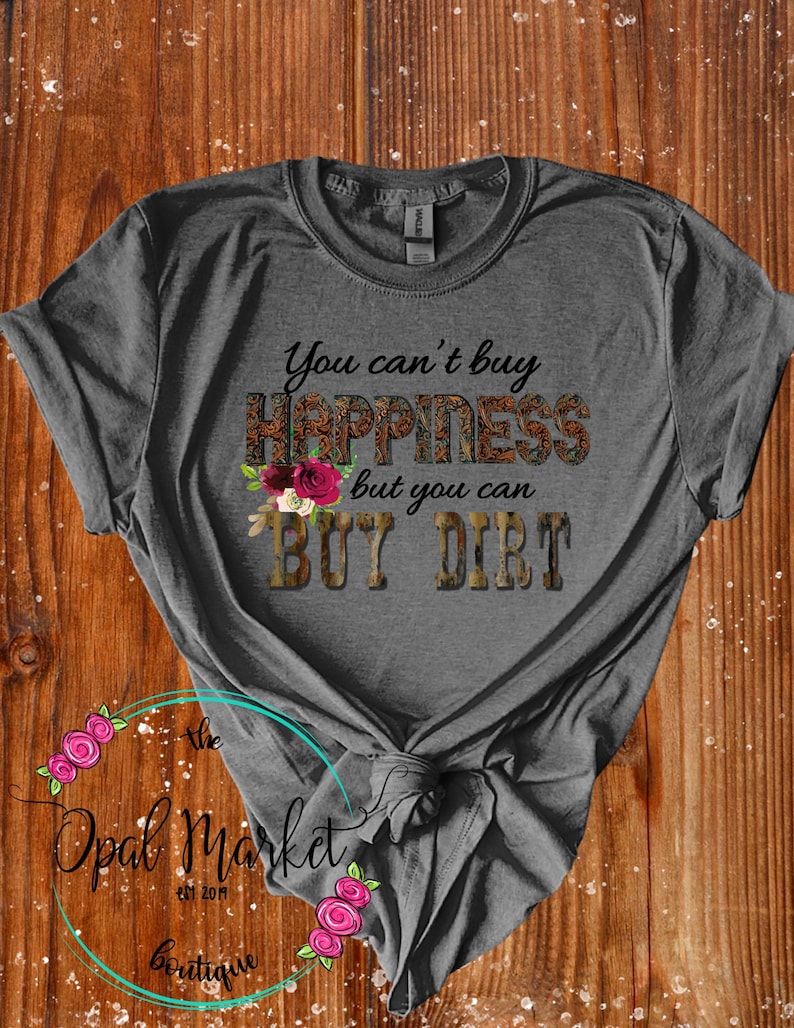 May include: Gray t-shirt with the text "You can't buy happiness but you can buy dirt" in brown and gold lettering with a floral design. The text is surrounded by a camouflage pattern.