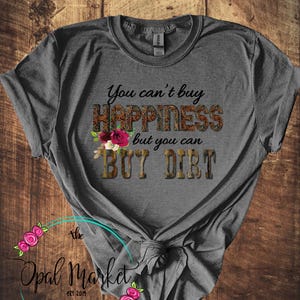 May include: Gray t-shirt with the text "You can't buy happiness but you can buy dirt" in black. The text is surrounded by a floral design and a camouflage pattern. The shirt is tied in a knot at the bottom.
