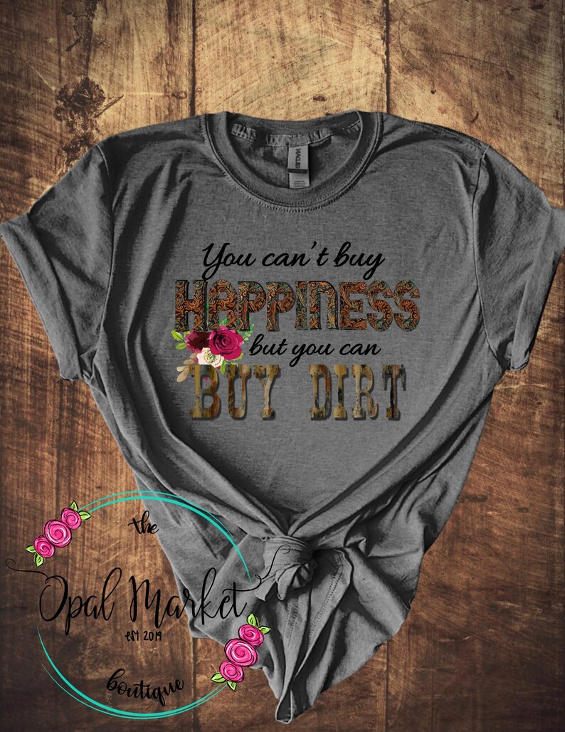 May include: Gray t-shirt with the text "You can't buy happiness but you can buy dirt" in black. The text is surrounded by a floral design and a camouflage pattern. The shirt is tied in a knot at the bottom.