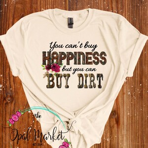 May include: A beige t-shirt with the text "You can't buy happiness but you can buy dirt" printed on it. The text is in a black font with a brown camouflage pattern and pink flowers. The text is centered on the shirt.