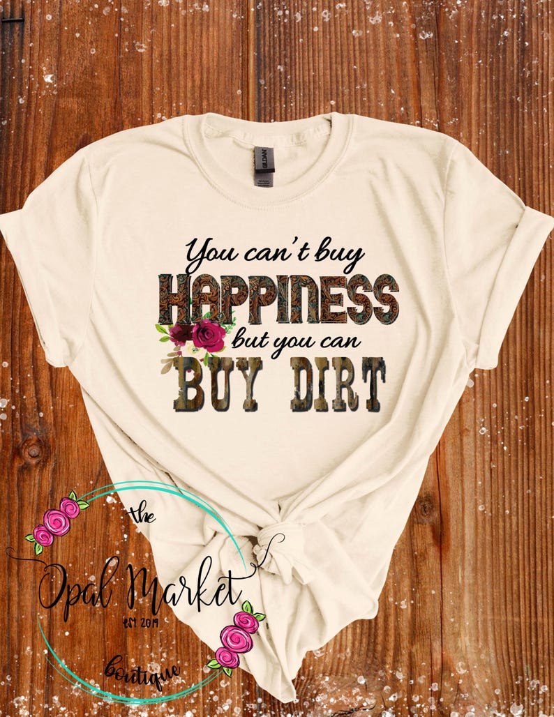 May include: A beige t-shirt with the text "You can't buy happiness but you can buy dirt" printed on it. The text is in a black font with a brown camouflage pattern and pink flowers. The text is centered on the shirt.