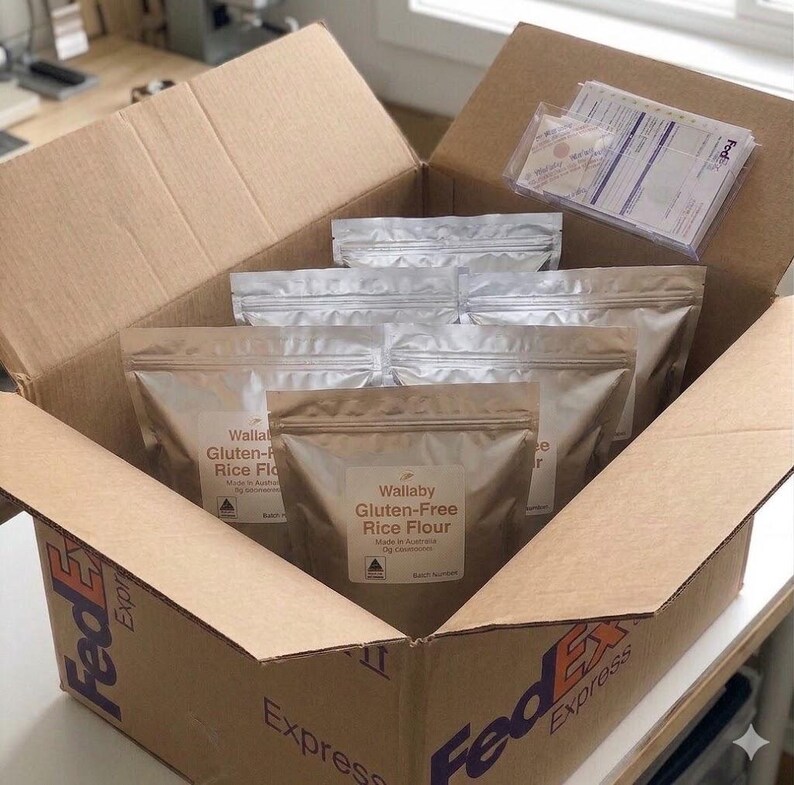 K&ouml;nnte beinhalten: Ein FedEx Express Karton mit mehreren Beuteln Wallaby Gluten-Free Reismehl. Die Beutel sind silber- und braunfarben und mit Etiketten versehen. Zus&auml;tzliche Beutel sind im Hintergrund sichtbar.