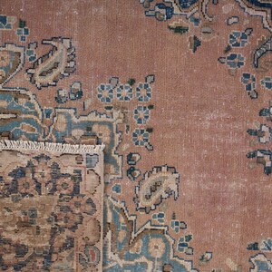 May include: Close-up of a vintage rug with a distressed, faded appearance. The rug features a central field of muted rose color, with intricate floral and paisley patterns in shades of blue, beige, and brown. The rug's fringe is visible at the bottom.