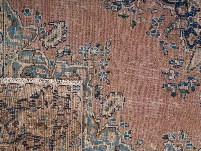 May include: Close-up of a vintage rug with a distressed, faded appearance. The rug features a central field of muted rose color, with intricate floral and paisley patterns in shades of blue, beige, and brown. The rug's fringe is visible at the bottom.
