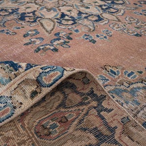 May include: Close-up of a vintage rug with a faded, distressed appearance. The rug features a central design in shades of peach, beige, and blue, with intricate floral and geometric patterns. The edge of the rug is folded over, revealing the detailed border.