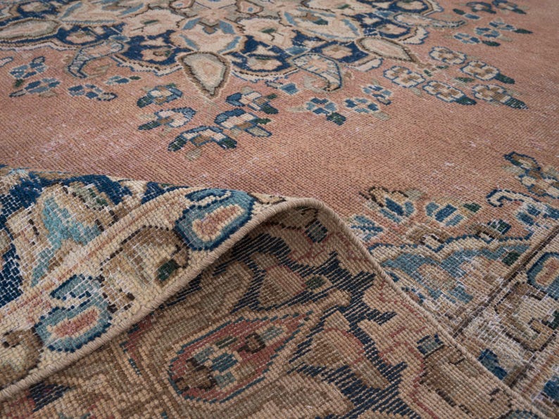 May include: Close-up of a vintage rug with a faded, distressed appearance. The rug features a central design in shades of peach, beige, and blue, with intricate floral and geometric patterns. The edge of the rug is folded over, revealing the detailed border.