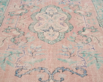 May include: A distressed, vintage-style rug with a faded pink and teal color palette. The rug features an ornate, symmetrical design with floral and geometric elements. The central medallion is cream-colored, and the rug has a distressed, worn appearance.