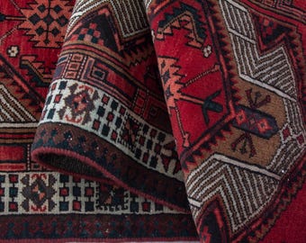 May include: Close-up of a rolled-up, intricately patterned rug. The rug features a rich color palette of red, brown, and cream, with geometric and floral designs. The edges are detailed with a dark border.