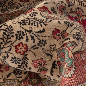 May include: Close-up of a beige, red, and teal vintage rug with an intricate floral pattern. The rug features a distressed look with visible wear and tear, adding to its antique charm. The design includes floral and leaf motifs.