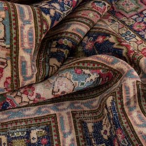 May include: Close-up of a folded, vintage rug with intricate patterns. The rug features a cream-colored central field with floral and geometric designs in shades of blue, red, and green. The borders are a darker blue with similar patterns.