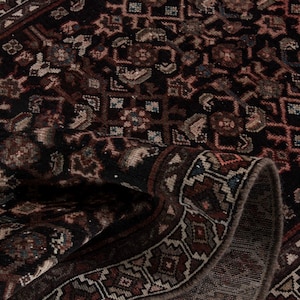 May include: Close-up of a dark, patterned rug with a rolled edge. The rug features a complex design with geometric shapes and floral motifs in shades of brown, red, and beige against a black background. The rug's texture is visible.