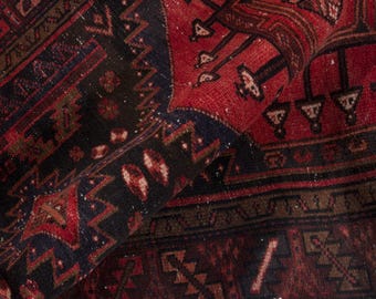 May include: Close-up of a rolled-up, vintage rug with a detailed, geometric pattern. The rug features a rich colour palette of red, black, and brown, with intricate designs and a textured appearance. The rug's design includes repeating shapes and borders.