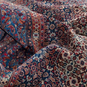 May include: Close-up of a detailed, folded rug with a traditional Persian design. The rug features a rich color palette of deep blues, reds, and cream, with intricate floral and geometric patterns. The texture and folds of the rug are visible.