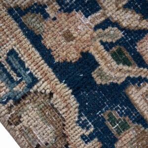 May include: Close-up of a section of a rug with a geometric pattern. The rug features a combination of colors, including navy blue, tan, and touches of green. The texture appears to be thick and woven, with visible knots and loops.