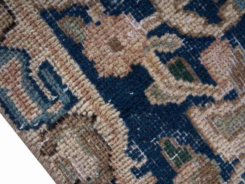 May include: Close-up of a section of a rug with a geometric pattern. The rug features a combination of colors, including navy blue, tan, and touches of green. The texture appears to be thick and woven, with visible knots and loops.