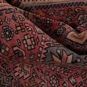 May include: Close-up of a folded, antique rug with a detailed, geometric pattern. The rug features a predominantly red colour with accents of black, yellow, and beige. The intricate design suggests a traditional, handcrafted textile.