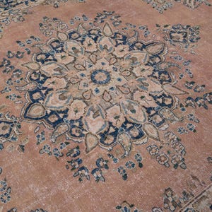 May include: A detailed close-up of a vintage rug with a central medallion design. The rug features a faded, distressed look with a color palette of muted rose, navy blue, and cream. The intricate floral patterns are visible.