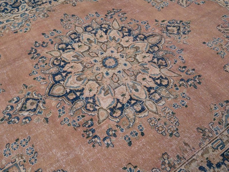 May include: A detailed close-up of a vintage rug with a central medallion design. The rug features a faded, distressed look with a color palette of muted rose, navy blue, and cream. The intricate floral patterns are visible.