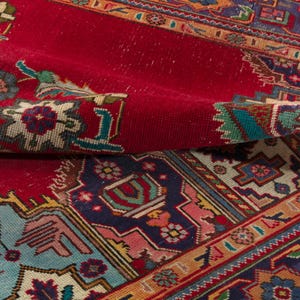 May include: Close-up of a vibrant red oriental rug with intricate geometric and floral patterns. The rug features a rich color palette including teal, navy, and gold accents. The rug is folded, showcasing its detailed design and texture.