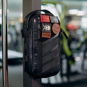 May include: Black tactical pouch with multiple patches. The patches include a red patch with white text, a skull and crossbones, and a brown patch with a mountain design. The pouch has a textured front and a zipper closure.