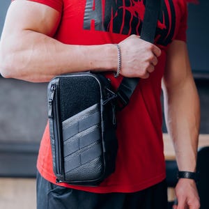 Op de afbeelding: Zwarte crossbodytas met een getextureerd voorpaneel en een vak met ritssluiting. De tas heeft een zwarte, verstelbare schouderriem. De persoon op de foto draagt een rood t-shirt met de zwarte tekst "ANIMAL".