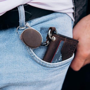 May include: A brown leather wallet with a silver key ring is in a light blue jeans pocket. A brown, circular key holder is attached to the jeans loop. The wallet has a card in it. The overall theme is accessories.
