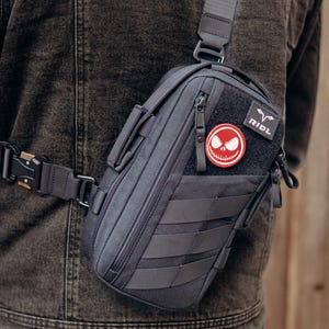 Peut inclure: Sacoche tactique gris foncé avec une sangle et une boucle noires. Le sac comporte un écusson avec un motif Jack Skellington et le mot "RIDL". Le sac a plusieurs compartiments et sangles.