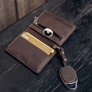 Könnte beinhalten: Eine braune Ledergeldbörse mit einem Reißverschlussfach, das einen silbernen Apple AirTag enthält, einer goldenen Visa-Kreditkarte in einem Schlitz und einem befestigten, einziehbaren Schlüsselhalter aus braunem Leder mit Metallclip.