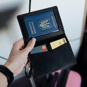 May include: Black leather passport holder with a clear window displaying a blue Ukrainian passport. The holder also contains a Mastercard and a Visa card. The holder is open, revealing the contents. The stitching is a light tan color.