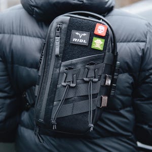 May include: Black leather crossbody bag with multiple compartments and straps. Features a zippered main compartment, a patch with the text "RIDL", and two patches with skull and crossbones designs. The bag is worn over the shoulder.