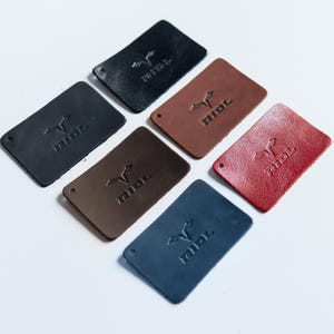 May include: Six rectangular leather tags in black, brown, tan, red, and blue, each featuring a debossed logo with the text RIDL and a small hole in one corner. The tags are arranged in a scattered pattern, showcasing varied textures and colors.