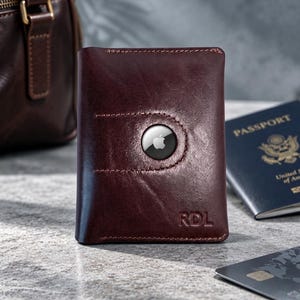 May include: A dark brown leather wallet featuring a circular cutout designed to hold an Apple AirTag. The wallet has visible stitching along the edges and the letters RDL embossed in the bottom right corner. The surface has a smooth texture.