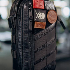 Op de afbeelding: Zwarte leren crossbodytas met meerdere patches. De tas heeft een verticale rits, een getextureerd oppervlak en een gestreept ontwerp aan de zijkant. Patches tonen een schedel, bergen en andere symbolen.