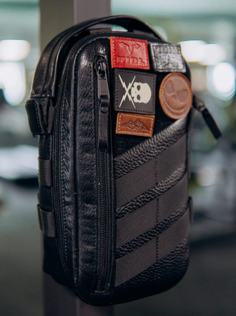 Op de afbeelding: Zwarte leren crossbodytas met meerdere patches. De tas heeft een verticale rits, een getextureerd oppervlak en een gestreept ontwerp aan de zijkant. Patches tonen een schedel, bergen en andere symbolen.