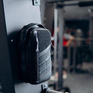 Op de afbeelding: Een zwarte, rechthoekige tas met een getextureerd voorpaneel en een ritssluiting. De tas is bevestigd aan een zwarte metalen structuur, waarschijnlijk in een sportschool. De tas heeft een handvat en een gewatteerd ontwerp.
