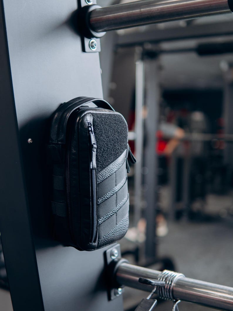 Op de afbeelding: Een zwarte, rechthoekige tas met een getextureerd voorpaneel en een ritssluiting. De tas is bevestigd aan een zwarte metalen structuur, waarschijnlijk in een sportschool. De tas heeft een handvat en een gewatteerd ontwerp.