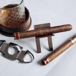 May include: A close-up of a cigar and accessories. A brown cigar rests on a metal stand. A second cigar with a red band lies next to a silver cigar cutter. A glass of amber liquid sits in the background.