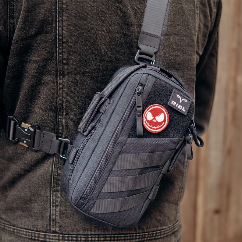 Op de afbeelding: Een donkergrijze tactische sling bag met een zwarte riem en gesp. De tas heeft een patch met een Jack Skellington-ontwerp en het woord "RIDL". De tas heeft meerdere compartimenten en riemen.