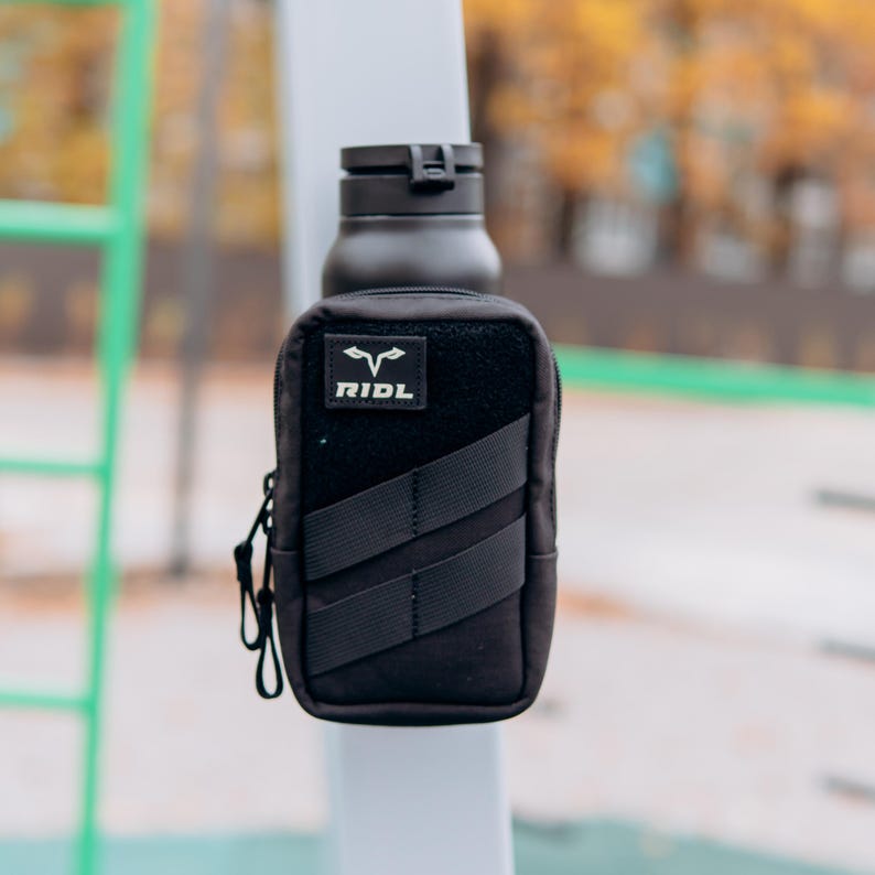Op de afbeelding: Zwarte waterfles met een zwart etui bevestigd. Het etui heeft een klittenband patch met de witte tekst "RIDL" en een logo. Twee zwarte bandjes zitten aan de voorkant. Een zwarte karabijnhaak is aan de zijkant bevestigd.